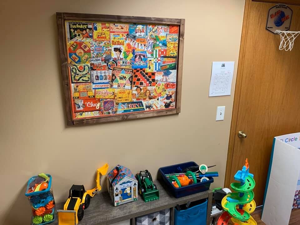 A framed vintage board-game puzzle above a shelf of trucks, marble runs and building toys, with a mini basketball hoop on the door