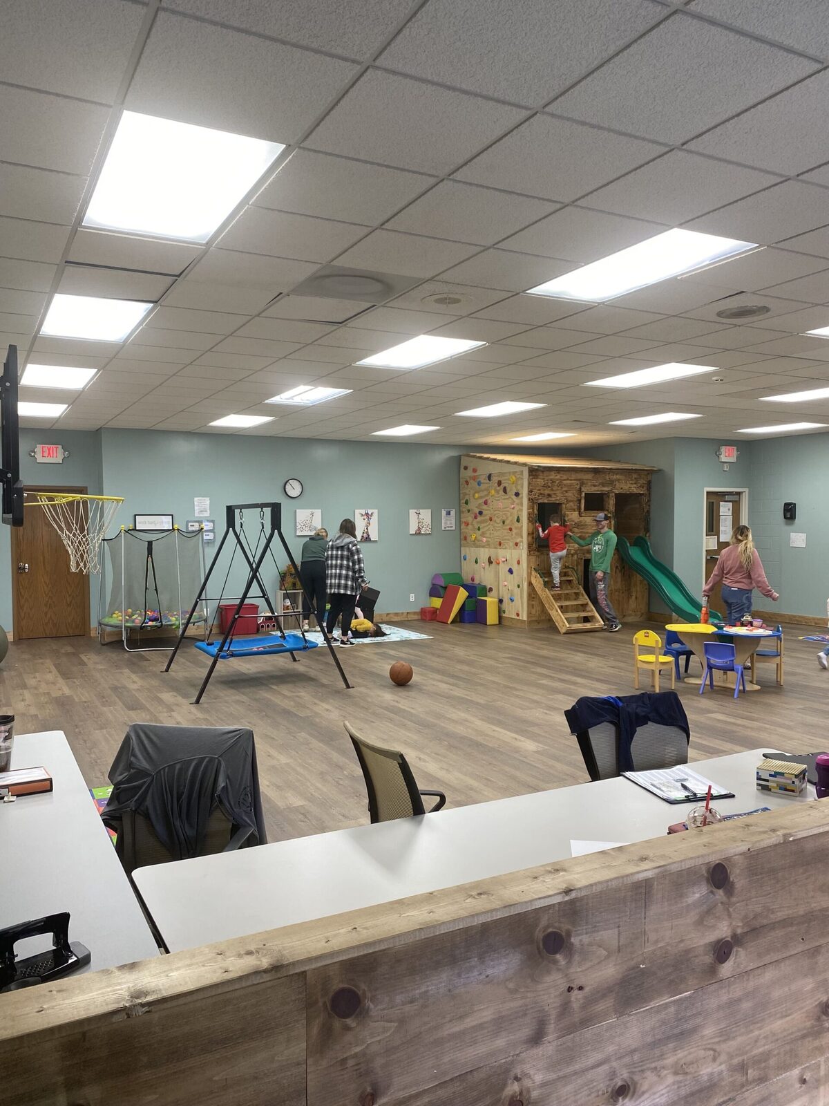 An open therapy gym seen from the front desk, with a basketball hoop, platform swing, trampoline and a wooden climbing tower with a green slide