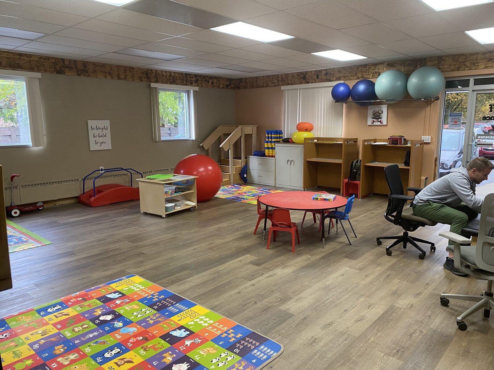 A therapy room with therapy balls racked along the wall, a wooden step platform, a rolling supply cart, an alphabet floor mat and a round activity table with small chairs