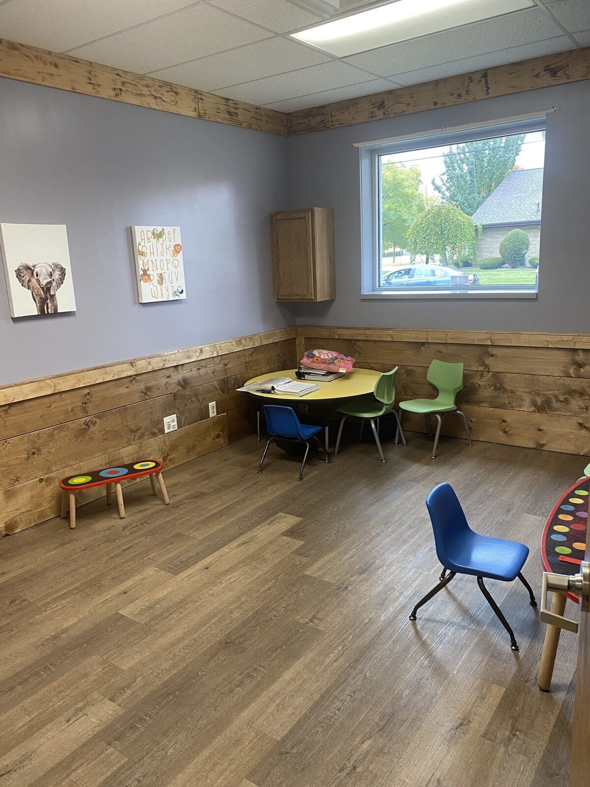 A quiet break-out room with plank wainscot, a round table and small chairs, a low balance bench and a window onto the street